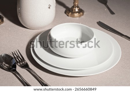 Close-up of elegant table setting with modern white ceramic plates and bowl, shiny cutlery, candle holder and soft beige tablecloth, minimalist fine dining atmosphere with bright directional lighting
