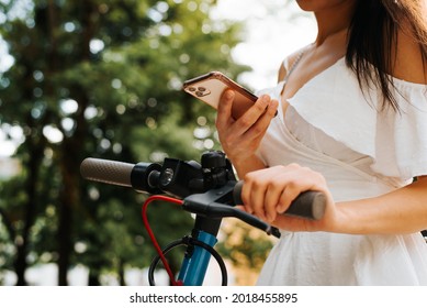 Close-up of elegant young woman in white summer dress renting a scooter using smartphone to pay online rent, outdoors. Electric scooter rental using mobile application. Selective focus on cell phone - Powered by Shutterstock