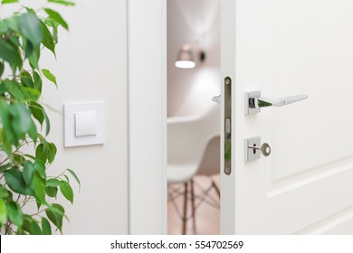 Close-up Elements Of The Interior Of The Apartment. Ajar White Door. Chrome Door Handle And Lock With Key. The Light Switch On The Wall