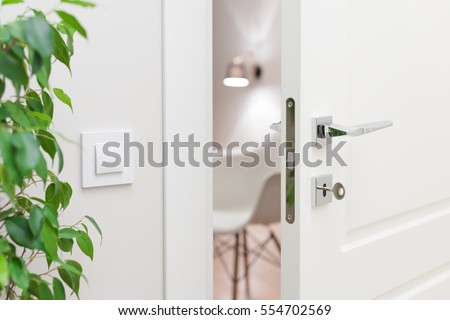 Close-up elements of the interior of the apartment. Ajar white door. Chrome door handle and lock with key. The light switch on the wall