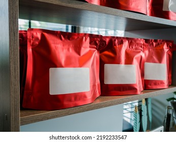 Closeup Empty Blank White Label Of Modern Red Coffee Bag Packaging. Mockup Template Sticker Label. Row Of Vivid Foil Paper Zipper Pouch Packaging For Coffee Bean Showing On Wooden Shelf.