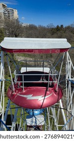close-up of empty red ferris wheel gondola with white canopy roof, lightning bolt decoration, metal safety rail, park trees and urban buildings visible in background, clear blue sky above