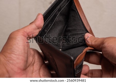 A close-up of an empty wallet held by a person's hands. The image symbolizes financial hardship, poverty, and lack of funds. The dark color of the wallet and the stark contrast with the empty interior