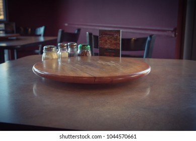 Close-up Empty Wooden Table With Rotating Serving Plate And Shaker Seasonings. Red Pepper Flakes, Parmesan, Black Pepper, Salt. Vintage Tone