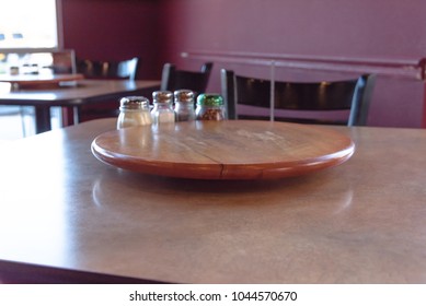 Close-up Empty Wooden Table With Rotating Serving Plate And Shaker Seasonings. Red Pepper Flakes, Parmesan, Black Pepper, Salt.