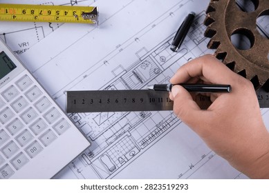 Close-up of an engineer or architect working on a building plan with ruler, calculator, tape measure, pen, and gear on a desk.