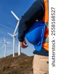 closeup Engineer wearing an orange safety vest holds a blue hardhat while walking near large wind turbines, symbolizing renewable energy and sustainable practices