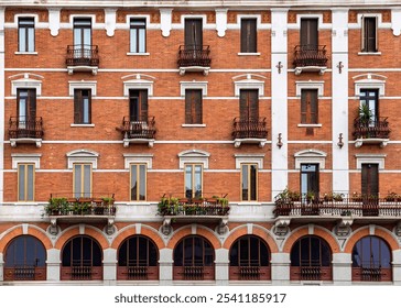 Close-up of a European apartment building with red brick facade, arched windows, wrought iron balconies, and potted plants on balconies.
 - Powered by Shutterstock