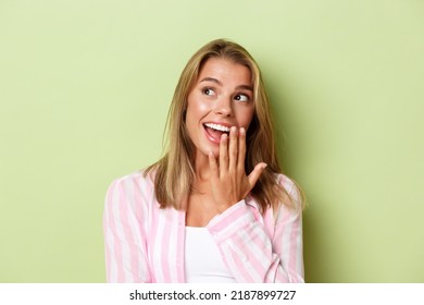 Close-up Of Excited Blond Girl In Pink Shirt, Looking At Upper Left Corner And Smiling Amused, Standing Over Green Background