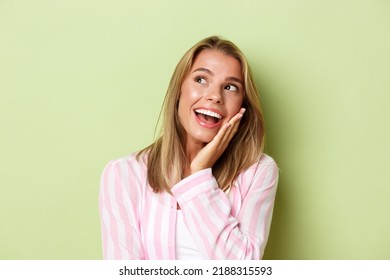 Close-up Of Excited Blond Girl In Pink Shirt, Looking At Upper Left Corner And Smiling Amused, Standing Over Green Background