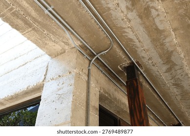 Close-up of exposed electrical conduit and cable tray mounted on a raw concrete beam in an industrial interior. Modern loft-style architecture with visible wiring system and structural details near window.