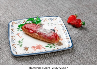 Close-up of an exquisite strawberry eclair with bright red glaze and powdered sugar. The dessert is garnished with fresh strawberries, a blueberry, and mint leaves, served on a decorative square plate