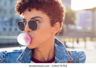 Closeup Face Of African Young Woman With Curly Hair Blowing Bubblegum Outdoor. Brazilian Girl Wearing Sunglasses And Denim Jacket Making Bubble Using Chewing Gum. Teenager Blowing Pink Bubble Gum.