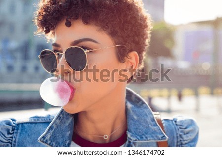 Closeup face of african young woman with curly hair blowing bubblegum outdoor. Brazilian girl wearing sunglasses and denim jacket making bubble using chewing gum. Teenager blowing pink bubble gum.