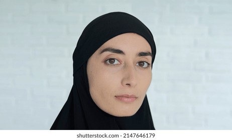 Close-up of the face of an Arab woman in a black hijab against a white brick wall, who turns her head and looks at the camera with an angry look. The recruitment of Muslim women by terrorists.