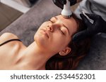 Closeup of face of beautiful relaxing girl undergoing hardware cosmetology procedure in spa. Cream for ultrasonic facial massage is applied to face of young woman using special nozzle in beauty salon.