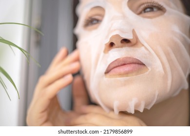 Closeup Face Beautiful Young Woman Applying Tissue Moisturizing Cooling Facial Mask For Skin Anti Aging Hydration. Domestic Female Enjoying Daily Routine Beauty Procedure Looking At Mirror In Bathroom