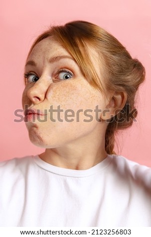 Close-up face of cute young girl crushed on glass isolated on light pink studio background in neon. Concept of human emotions, facial expression, youth. Model leaning against glass