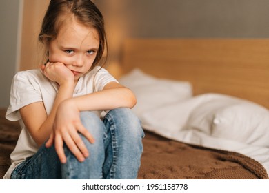 Close-up face of frowning beautiful little girl with wet eyes from tears sits on bed in bedroom and looking at camera. Depressed child feeling pressure, thinking of kid problems.