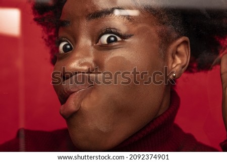 Close-up face of funny dark skinned young girl crushed on glass isolated on dark red studio background. Concept of human emotions, facial expression, youth. Model leaning against glass