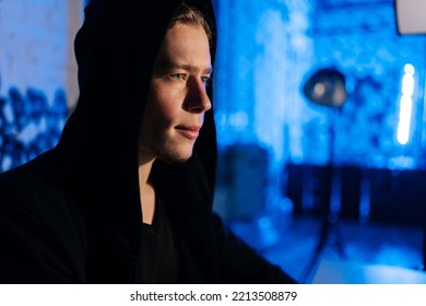 Close-up Face Of Handsome Wanted Hacker Man In Sweatshirt With Hood Engaged In Hacking Into Security Systems, Sitting In Dark Room With Blue Neon Lights. Network Hackers Steal Computer System Data.