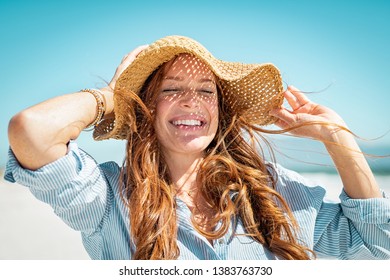 Closeup face of mature woman wearing straw hat enjoying the sun at beach. Happy woman smiling during summer vacation at sea.Beautiful lady relaxing at beach while holding large brim for the wind.
