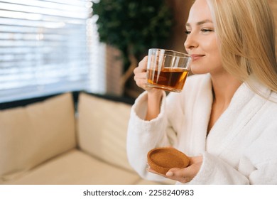 Close-up face of smiling young woman in white bathrobe enjoying drinking hot healthy tea after spa procedures. Relaxed blonde female resting with herbal drink sitting in sofa at spa salon. - Powered by Shutterstock