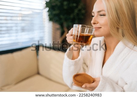 Close-up face of smiling young woman in white bathrobe enjoying drinking hot healthy tea after spa procedures. Relaxed blonde female resting with herbal drink sitting in sofa at spa salon.