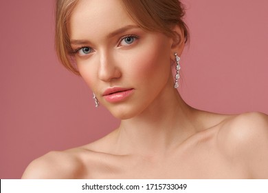 Closeup face studio portait of young elegant blonde caucasian woman with natural makeup and hairstyle wearing jewelry, earrings and posing against pink background. Fashion model