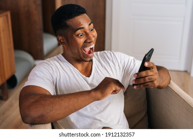 Close-up Face Of Surprised African-American Man Receiving Good News Using Mobile Phone At Home. Shocked Male With Open Mouth Enjoying Smartphone Communication Looking With Amazement At Message On Cell