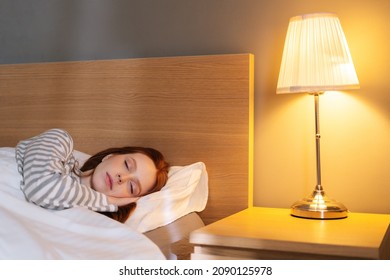 Close-up face of tired young woman sleeping well in bed hugging soft white pillow at home, bedside lamp lighting with warm yellow light. Cute lady resting enjoying fresh soft bedding linen in bedroom