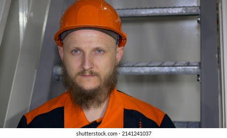 Close-up Of The Face Of A Worker In A Hard Hat, Working In A Factory In A Heavy Industry Or In The Energy Sector, As Well As In Mining. Bearded Man With Blue Eyes In Overalls Looking At The Camera