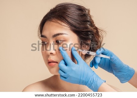 Closeup face young asian woman is getting filler under her eye for anti age wrinkle by a beautician with a syringe in hands. Cosmetic rejuvenating facial treatment concept. Aesthetic medical.