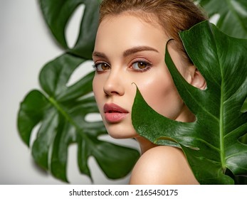 Closeup face of young beautiful woman with a healthy clean skin. Beautiful white girl with big green leaves. Beauty and spa treatment concept. Pretty woman with natural makeup and plant near face