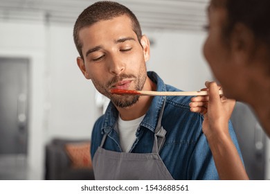 Closeup Face Of Young Man Blowing On Spoon To Taste Tomato Sauce While Woman Cooking In Kitchen. Guy Tasting Food For Dinner. Girl Offering To Her Boyfriend A Wooden Spoon With Sauce To Taste.