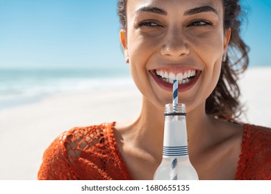 Closeup face of young woman drinking fresh sparkling water from a glass bottle at beach. Portrait of beautiful carefree girl drinking soft drink with straw during summer. Smiling girl staying hydrated