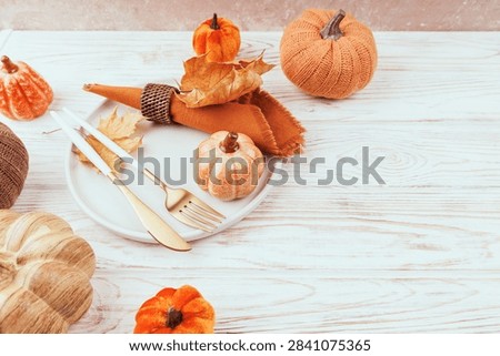 Close-up of fall table decor with ceramic and velvet pumpkins, orange napkin, maple leaves and golden cutlery on light wood surface