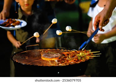 Close-up of Family grills skewers and roasts marshmallows over an open flame during a nighttime barbecue. The scene captures the cozy, festive atmosphere of outdoor cooking while camping - Powered by Shutterstock