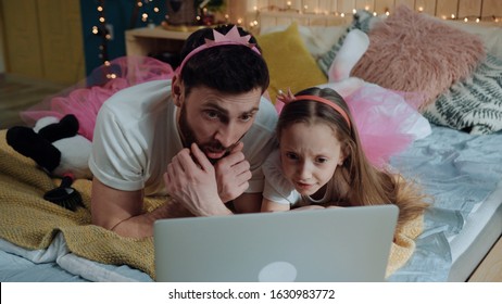Closeup family portrait young dad and girl dressed like fairies watching scary cartoons together on laptop computer lying on bed at home. Birthday party celebration.