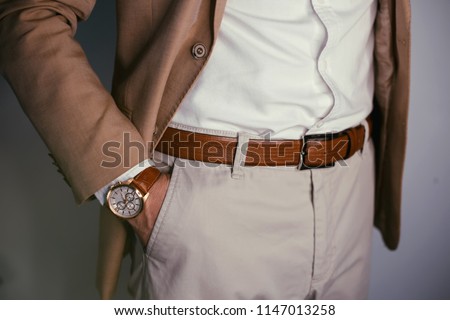Closeup fashion image of luxury watch on wrist of man.body detail of a business man.Man's hand in beige pants pocket closeup at white background.Man wearing brown  jacket and white shirt.Not isolated