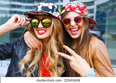 Closeup fashion lifestyle portrait of two pretty best friends girls, wearing bright swag style floral hats, mirrored sunglasses, having fun and make crazy funny faces. Two sisters posing on party.