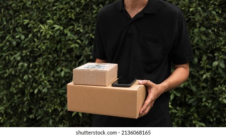 Closeup Fast And Express Delivery Man Standing In Black Shirt Carrying Cardboard Boxes Or Parcel With Green Leaves Nature Garden Background And Copy Space. Environmentally Friendly Business Concept