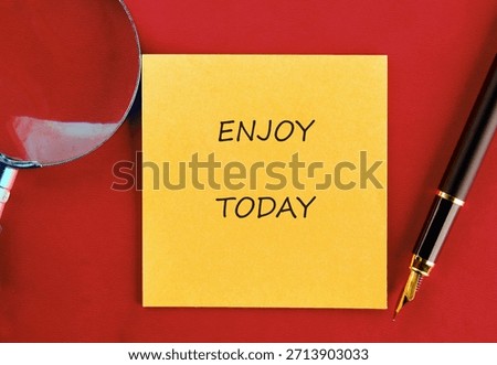 Close-up features a yellow sticky note with the phrase ENJOY TODAY, a magnifying glass, and a sophisticated fountain pen displayed against a vibrant red surface.