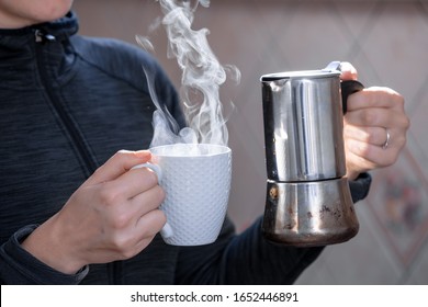 Closeup Of Female Adult Hands Holding White Cup In Right Hand, Metal Coffee Pot In Left Hand, Coffee Smoke Is Steaming Up