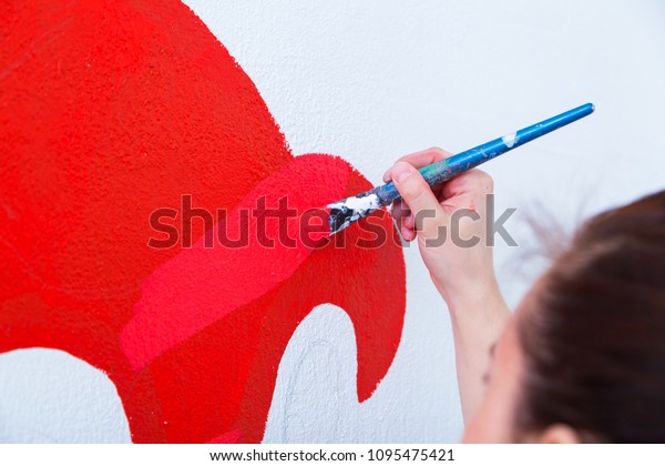 Close-up of a female artist in black and white shirt\
draws a red car on a wall with a pencil on a white wall in a\
children\'s room