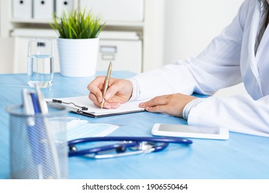 Close-up of female doctor filling medical form at clipboard. Therapist working at desk in office. Examination and diagnosis in modern clinic. Doctor workplace with green plant in pot and stethoscope.