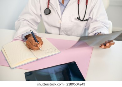 Close-up of female doctor making notes at table with x-ray image. African American general practitioner sitting at table and holding radiological picture. Pulmonology concept