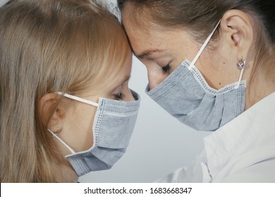 Close-up Of Female Doctor Wearing Medical Mask Sitting Face To Face With Little Blonde Girl In Medical Mask. The Doctor Calms The Baby And Hugs. Mom Consoles Scared Child