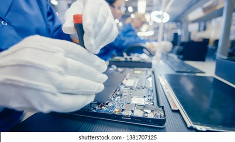 Close-Up Of A Female Electronics Factory Worker In Blue Work Coat Assembling Laptop's Motherboard With A Screwdriver. High Tech Factory Facility With Multiple Employees.