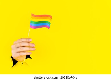 Closeup female hand is holding LGBTQ community rainbow flag color in torn hole of yellow background. LGBT gay pride parade concept. Love has no gender. Tolerance, freedom. Pansexual movement. 